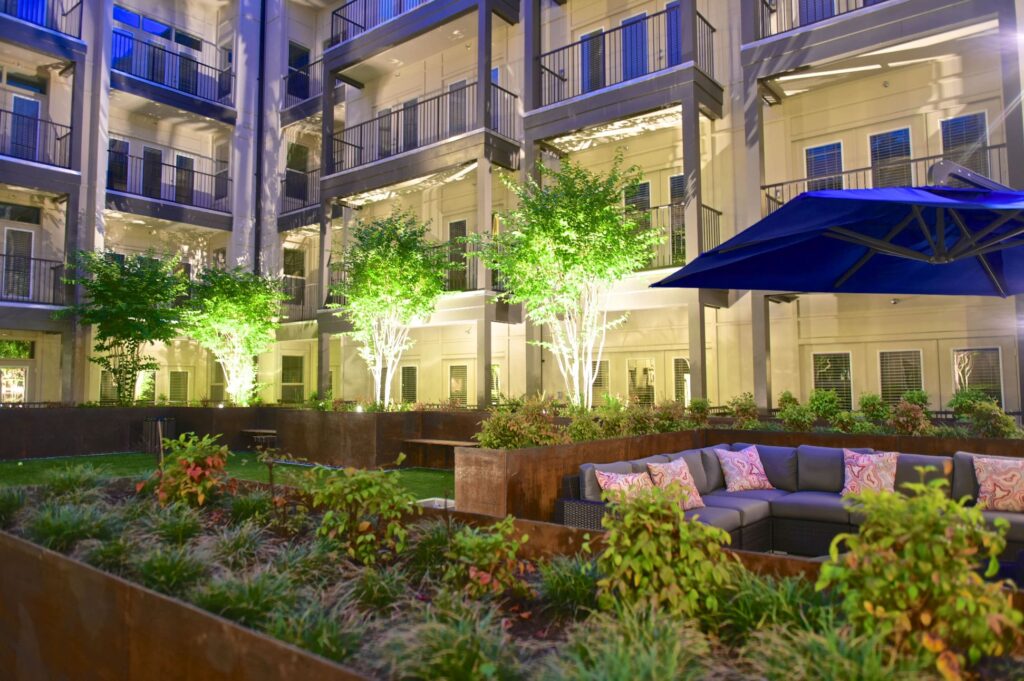 Courtyard at night with raised planters, lighting, patio lounge seating, and umbrella covering