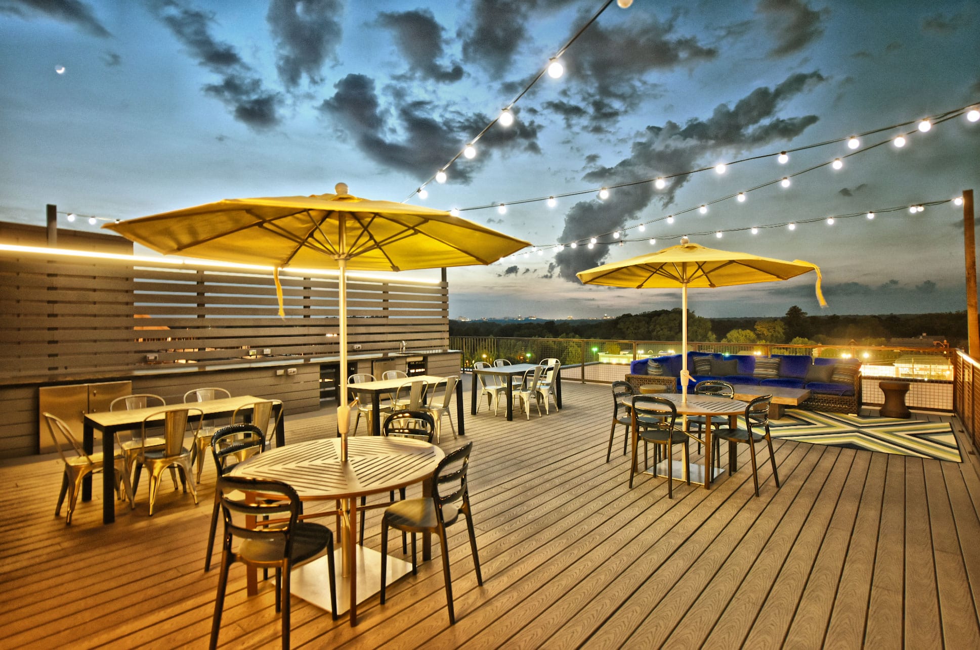 Rooftop amenity area with wood deck flooring, dining tables and chairs, umbrella coverings, patio lounge seating, overhead string lighting, and an outdoor kitchen wall