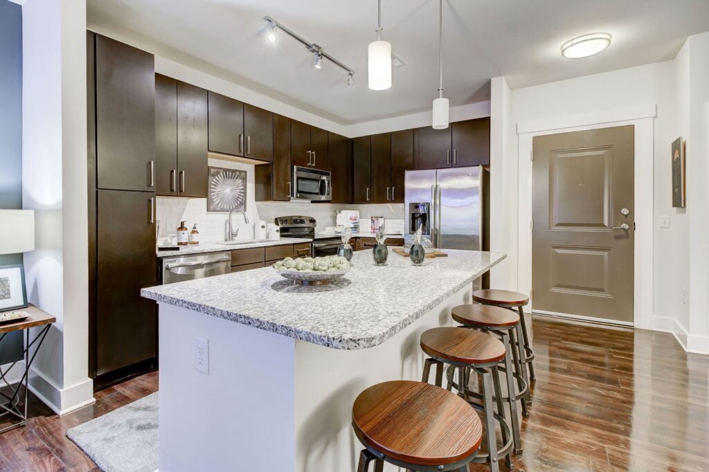 Apartment kitchen with large island and barstool seating, wood flooring, dark cabinetry, light granite countertops, white subway tile backsplash, stainless steel appliances, and pendant and track lighting