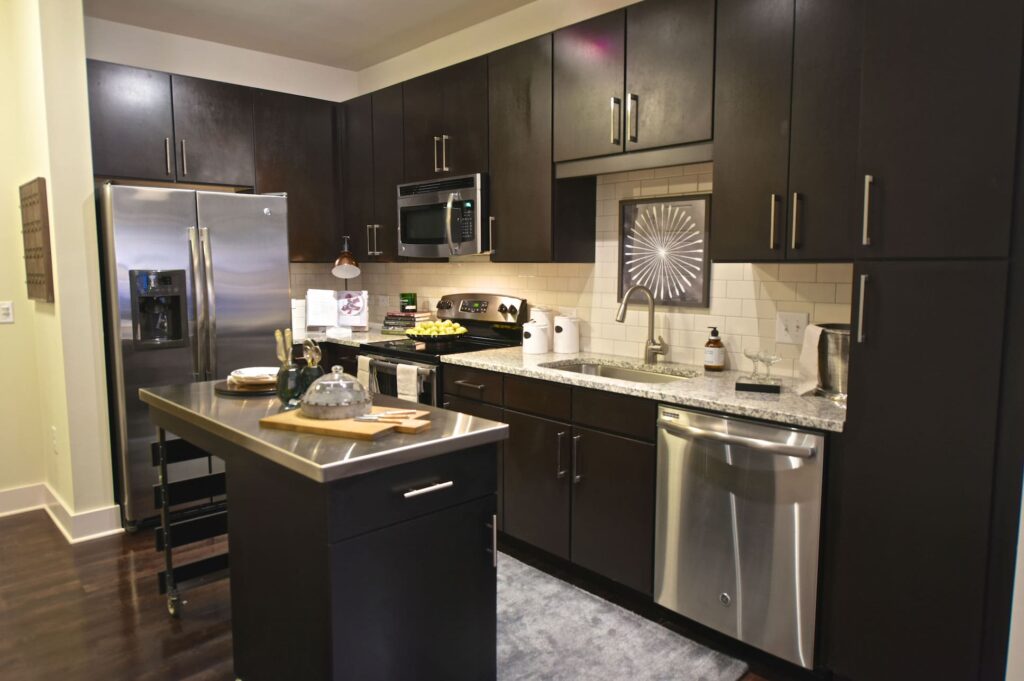 Apartment L-shaped kitchen with island on wheels, wood flooring, dark cabinetry, light granite and stainless steel countertops, white subway tile backsplash, and stainless steel appliances