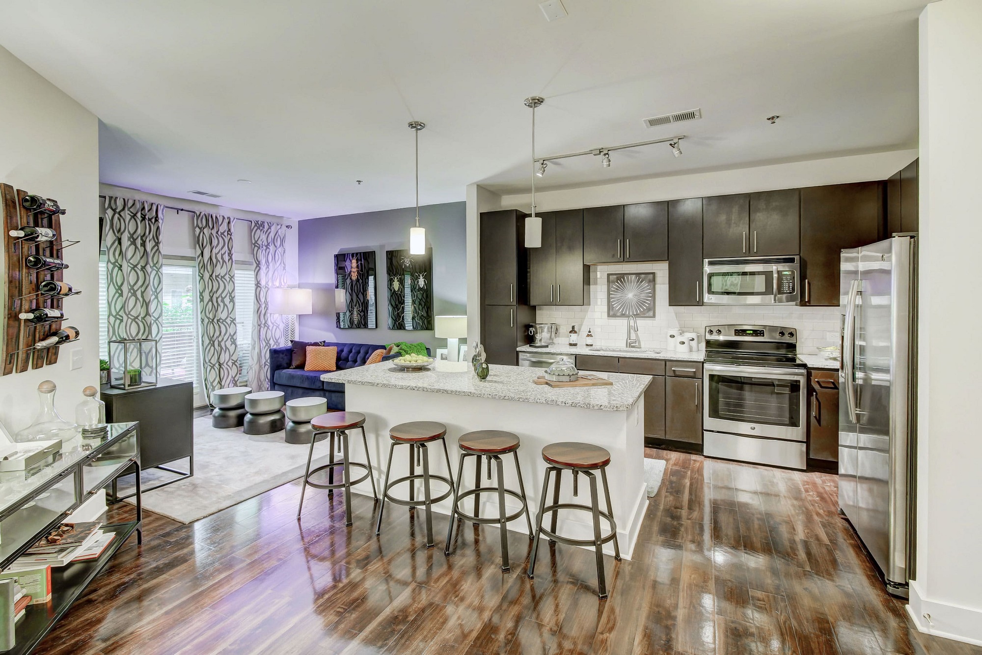 Furnished apartment with an open concept living room and kitchen including wood floors throughout, pendant and track lighting, dark cabinetry, stainless steel appliances, granite countertops, and an island with barstool seating
