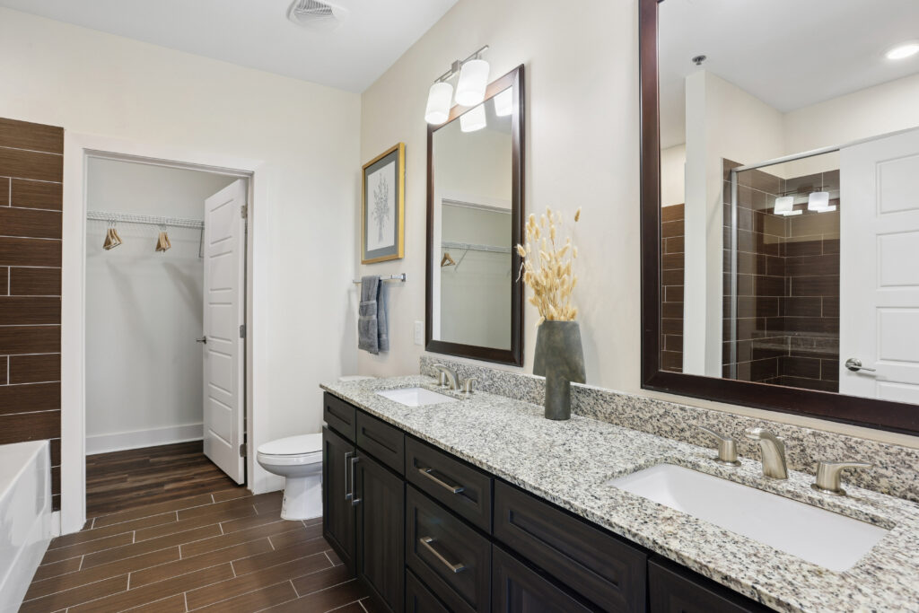 Modern bathroom with double sinks, granite countertop, dark wood cabinets, large mirrors, wall art, vase with dried plants, toilet, walk-in closet, and a glass-enclosed shower, featuring brown tile flooring and white walls.