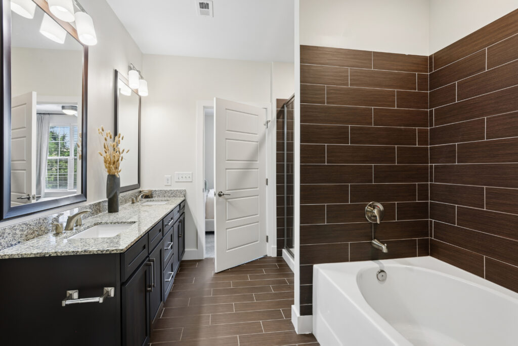 Modern bathroom with a double sink vanity, granite countertop, and dark cabinets. There is a white bathtub with brown tile surround and matching floor tiles, and a door open to a walk-in shower.
