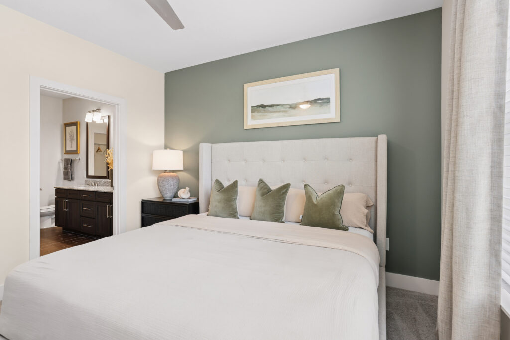 A modern bedroom with a beige upholstered bed, four green pillows, a black nightstand with a lamp, and a green accent wall. A bathroom with a double vanity is visible through an open doorway.