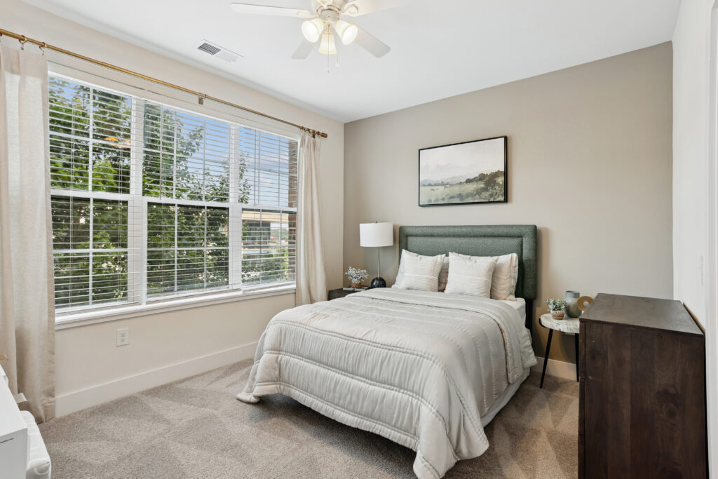 A bright bedroom with a large window, beige walls, a ceiling fan, a bed with a gray headboard, light bedding, a nightstand with a lamp, a dresser, and a landscape painting above the bed.