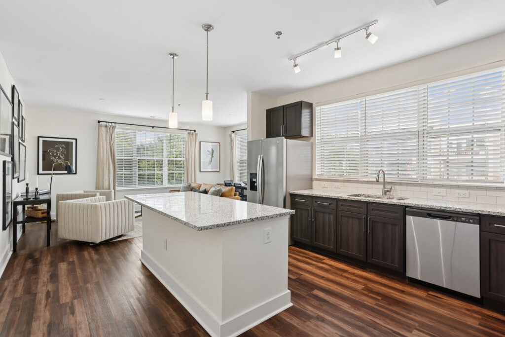 Modern kitchen with dark cabinets, granite island, stainless steel appliances, and hardwood floors, open to a bright living area with large windows and neutral furniture.