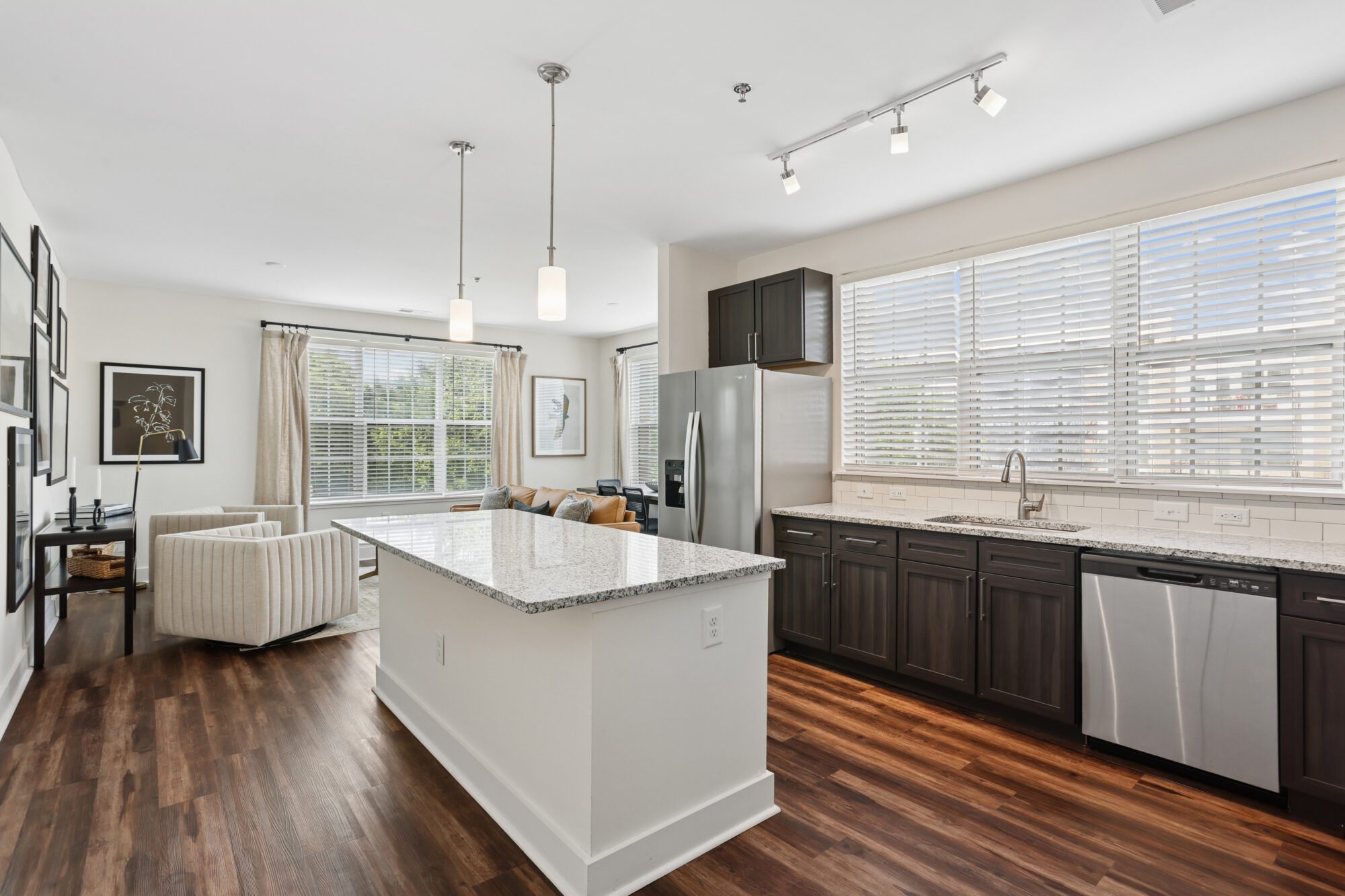 Modern kitchen with dark cabinets, granite island, stainless steel appliances, and hardwood floors, open to a bright living area with large windows and neutral furniture.
