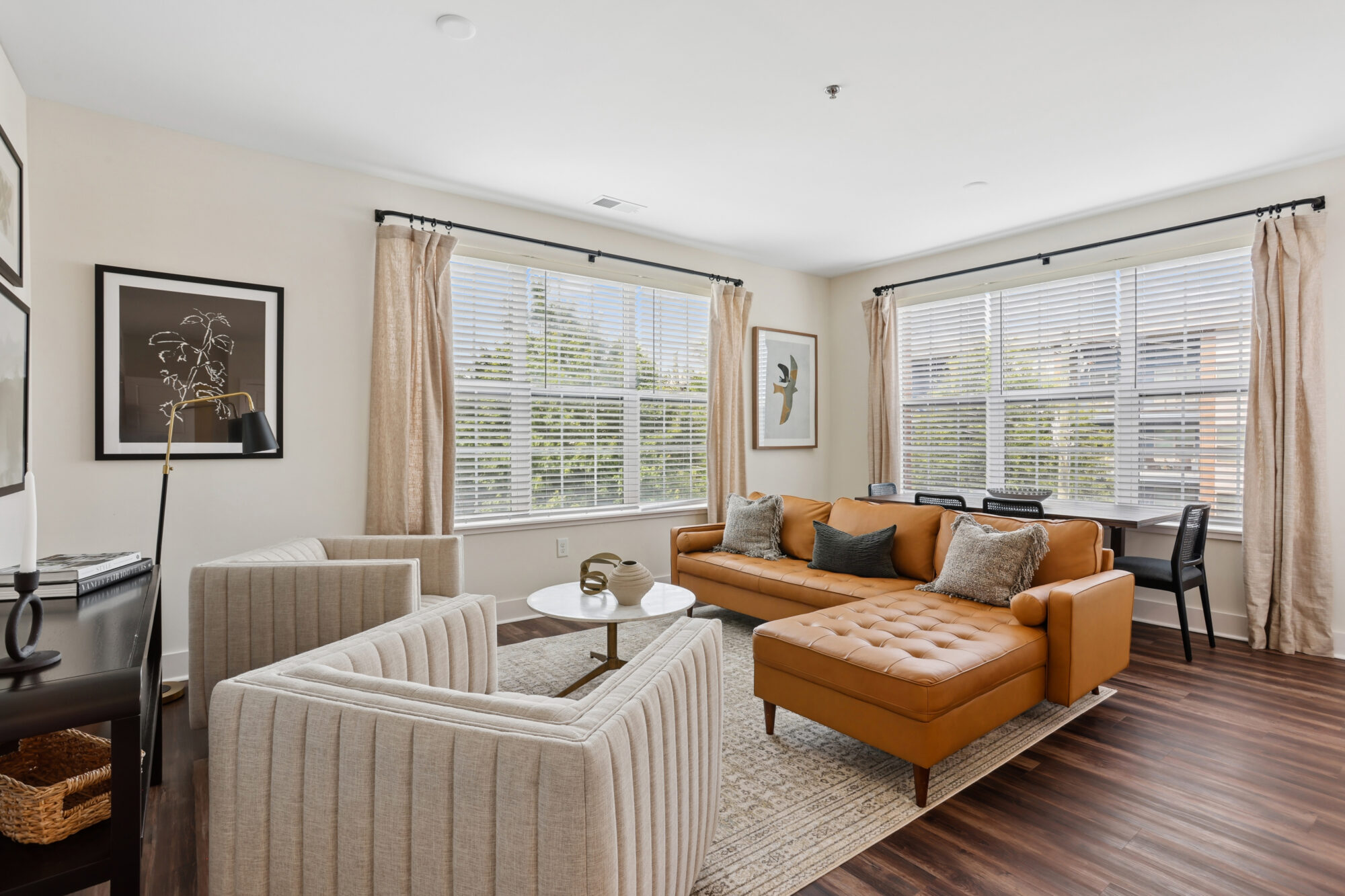A modern living room with a tan tufted sofa, two beige armchairs, a coffee table, large windows with beige curtains, framed art on white walls, a desk by the window, and dark wood flooring.