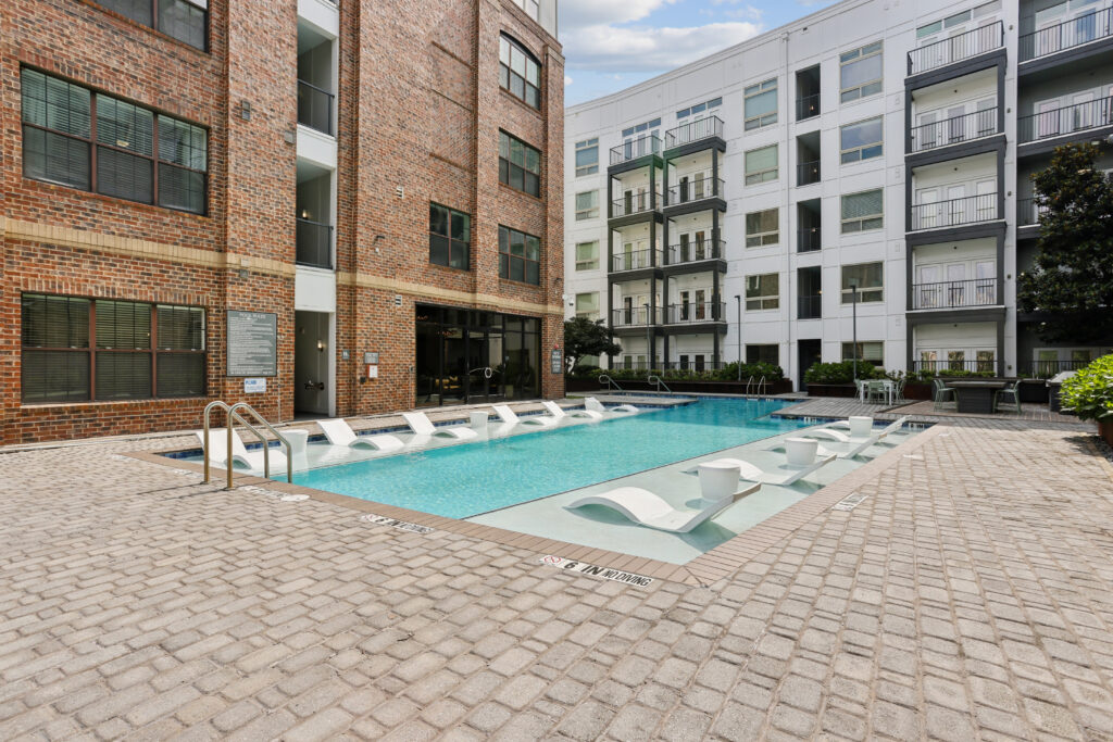 Outdoor swimming pool surrounded by lounge chairs and brick paving, with modern apartment buildings in the background. The pool has in-water loungers and is set in a well-maintained residential courtyard.