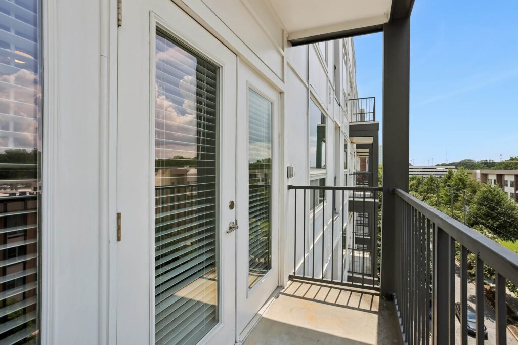 Small balcony with black railing, glass door with white blinds, overlooking a tree-filled area and other apartment buildings on a sunny day.