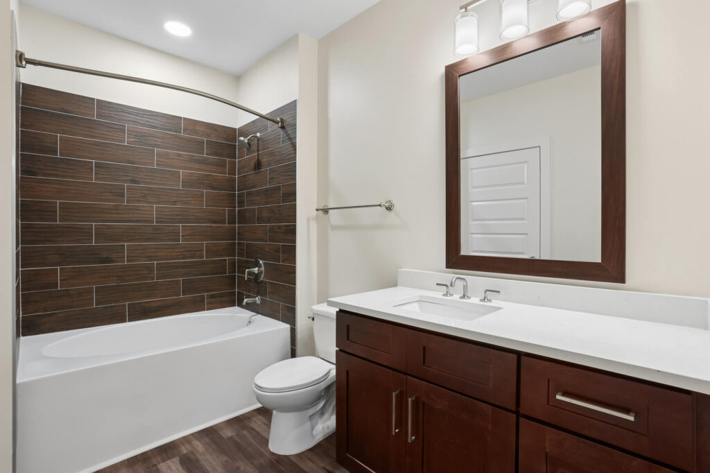 Modern bathroom with a white bathtub, dark wood-style tile wall, white toilet, brown vanity with white countertop, large mirror, and bright overhead lights.
