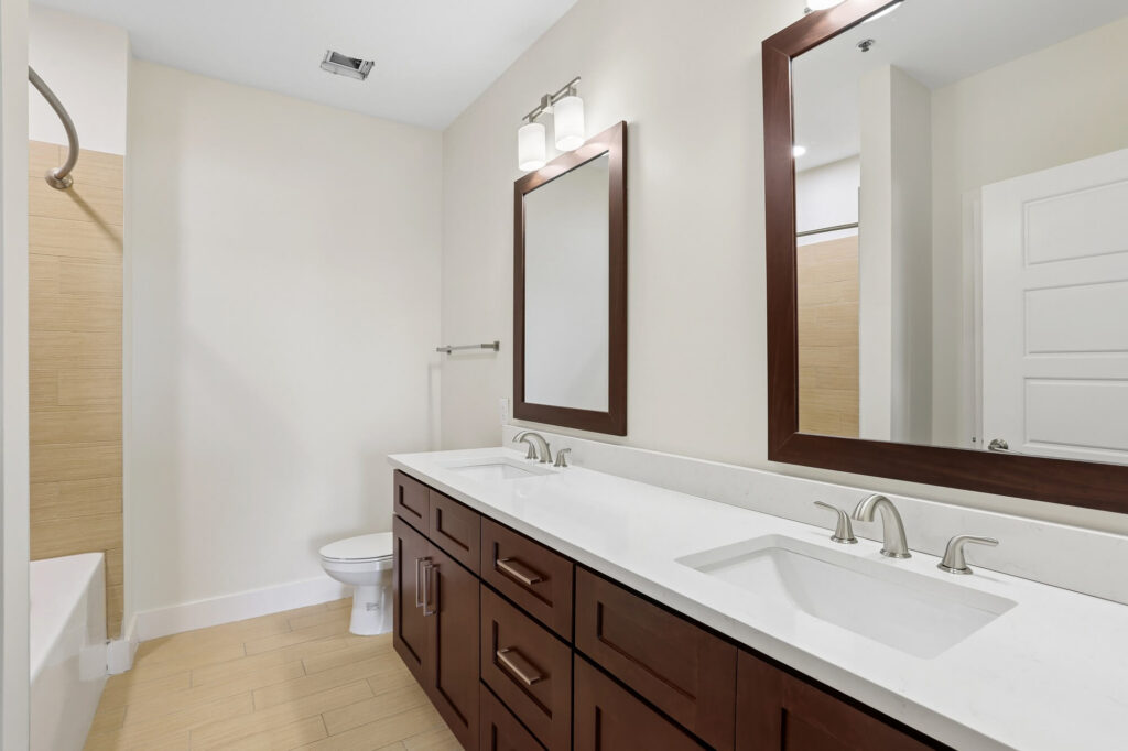 Modern bathroom with double sinks, dark wood cabinets, two mirrors, white countertops, toilet, and bathtub with a curved shower rod. Neutral walls and tile flooring create a clean, minimalist look.