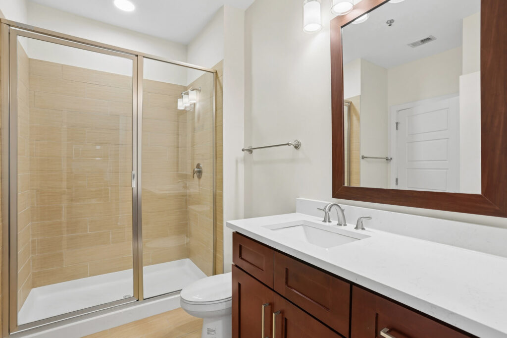 Modern bathroom with glass shower, wooden vanity with white countertop, rectangular mirror, and neutral walls. Bright lighting and chrome fixtures enhance the clean, contemporary look.
