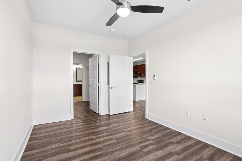 Empty room with white walls, dark wood flooring, ceiling fan with light, and two open white doors leading to a bathroom and kitchen area. Electrical outlets are visible on the right wall.