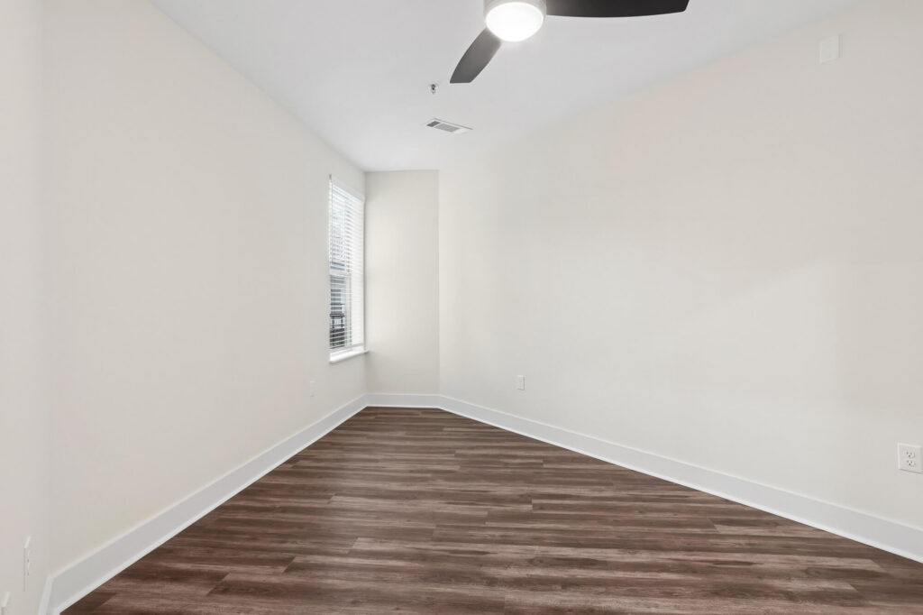 Empty room with white walls, dark wood flooring, a window with blinds on the left wall, ceiling fan with light, and white baseboards. The room has a slight angle and is brightly lit.