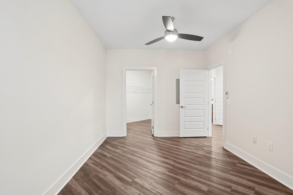 Empty room with light-colored walls, wood-patterned flooring, a ceiling fan, and two white doors—one open to a closet and another to a hallway. The room is well-lit and minimalistic.