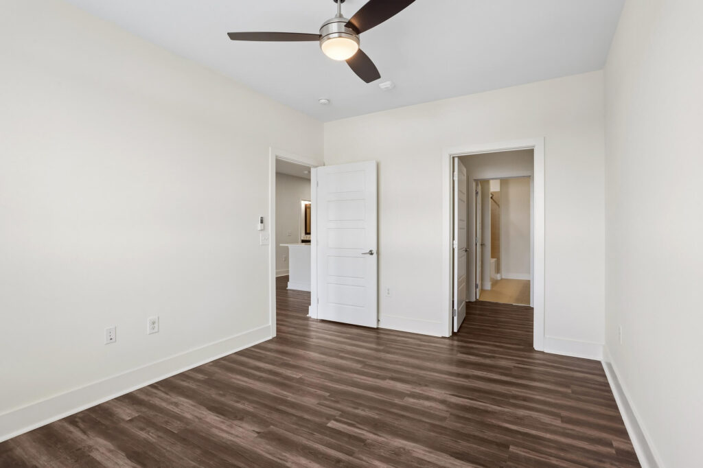 Empty room with light walls, dark wood flooring, a ceiling fan with light, and two open white doors leading to other rooms. Electrical outlets are visible on one wall.