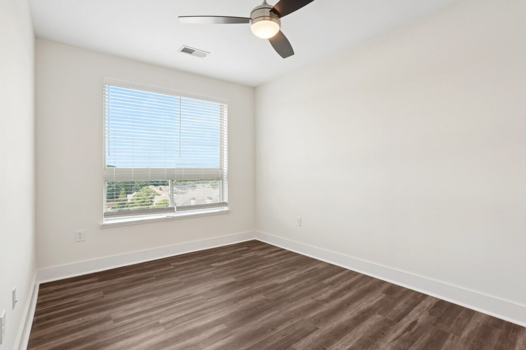 Empty room with light beige walls, a window with blinds letting in natural light, dark wood flooring, and a ceiling fan with a light.