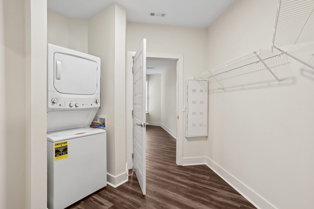 A laundry room with a stacked washer and dryer, an open white door, wire shelving on the right wall, and wood-look flooring. Walls are light-colored and the space is clean and minimal.