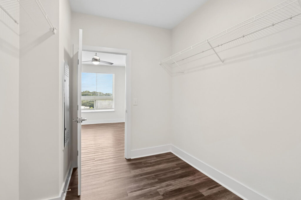Empty walk-in closet with white walls, wire shelving on the right, and wood-patterned flooring. An open door leads to a bright room with large windows, allowing natural light to fill the space.