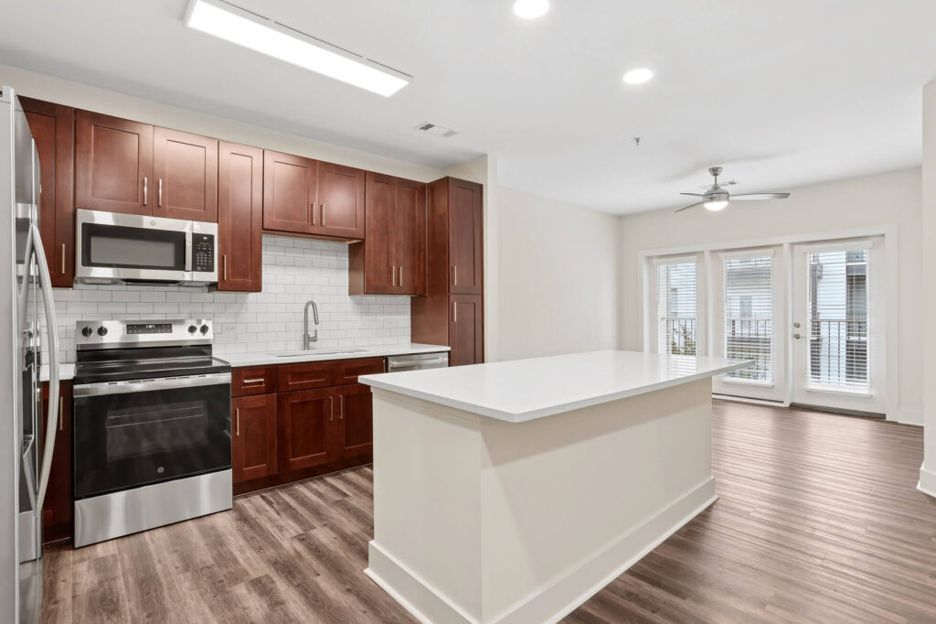Modern kitchen with dark wood cabinets, stainless steel appliances, white subway tile backsplash, large white island, and wood flooring. Open layout leads to a bright living area with French doors and ceiling fan.