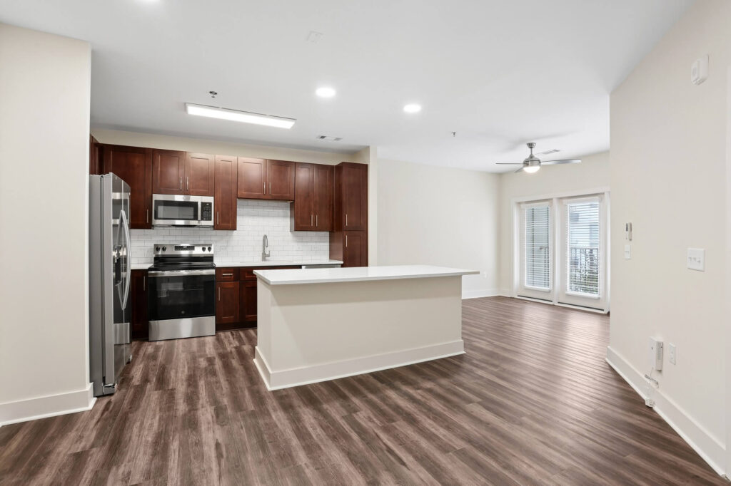 Modern kitchen with dark wood cabinets, stainless steel appliances, white countertops, and an island. The space has wood flooring and opens to a bright living area with large windows and a ceiling fan.