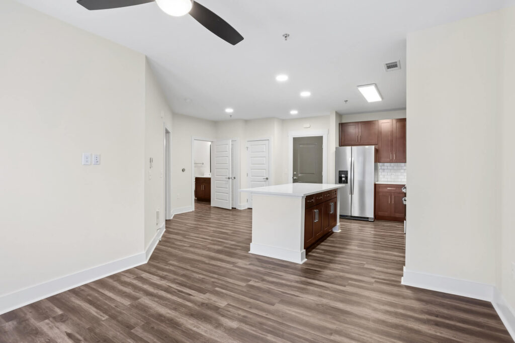 Modern kitchen with stainless steel appliances, wood cabinets, a center island, and wood-style flooring. White walls, ceiling fan, and doors leading to other rooms are visible. Bright, neutral, and spacious interior.