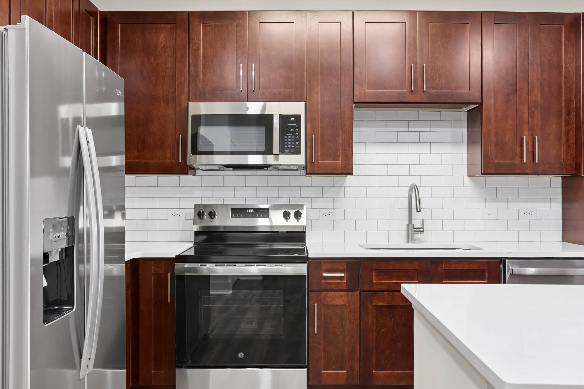 Modern kitchen with stainless steel appliances, dark wooden cabinets, white subway tile backsplash, electric stove, microwave, refrigerator, sink, and white countertops.