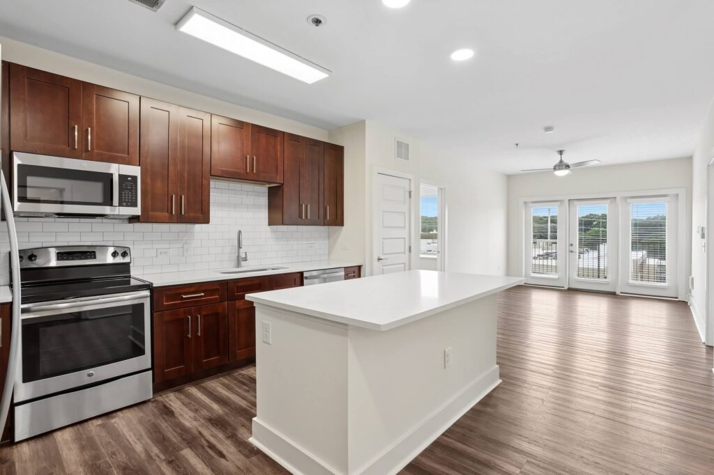 Modern kitchen with dark wood cabinets, stainless steel appliances, a white subway tile backsplash, and a large white island. The open layout leads to a bright living area with large windows and wood flooring.