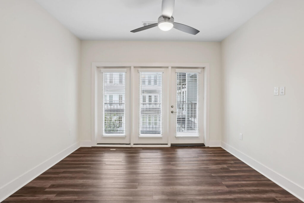 A bright, empty room with white walls, wood flooring, a ceiling fan, and double glass doors with blinds that open to a balcony.