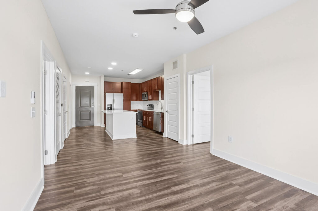Modern apartment interior with wood flooring, ceiling fan, open kitchen with dark wood cabinets, stainless steel appliances, and white countertops. Multiple doors lead to other rooms. Neutral-colored walls throughout.