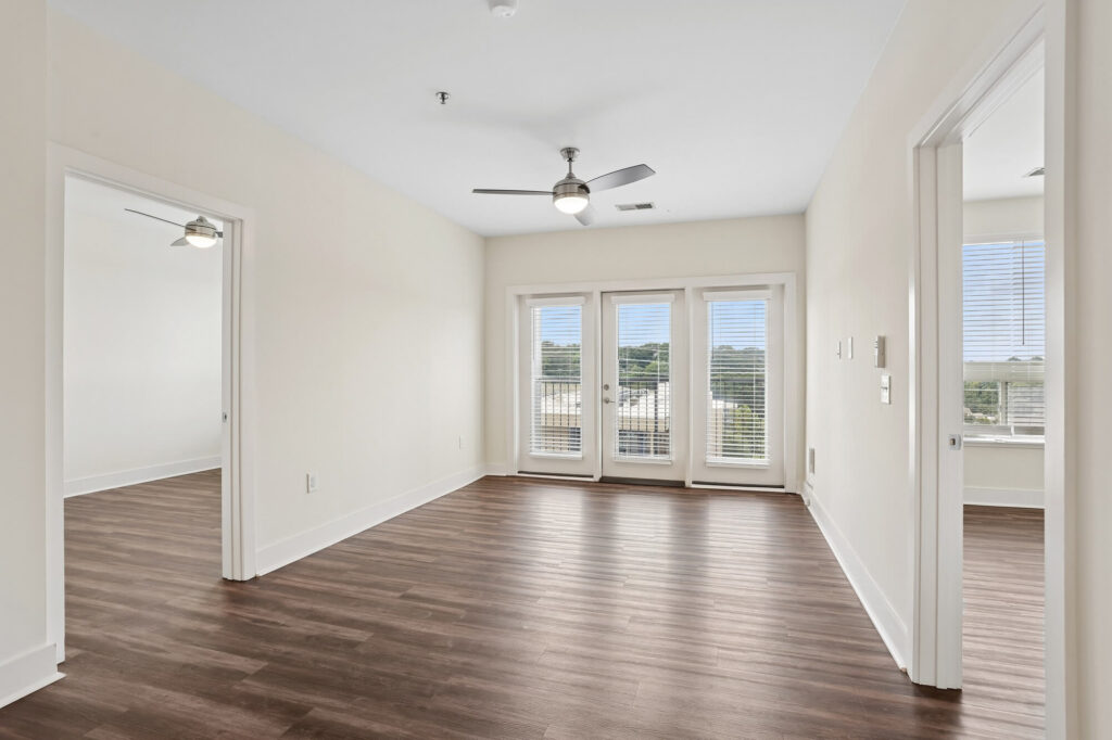 Bright, unfurnished living room with wood floors, two ceiling fans, and large windows with doors leading to a balcony. Two doorways on either side open to adjoining rooms with similar flooring and windows.