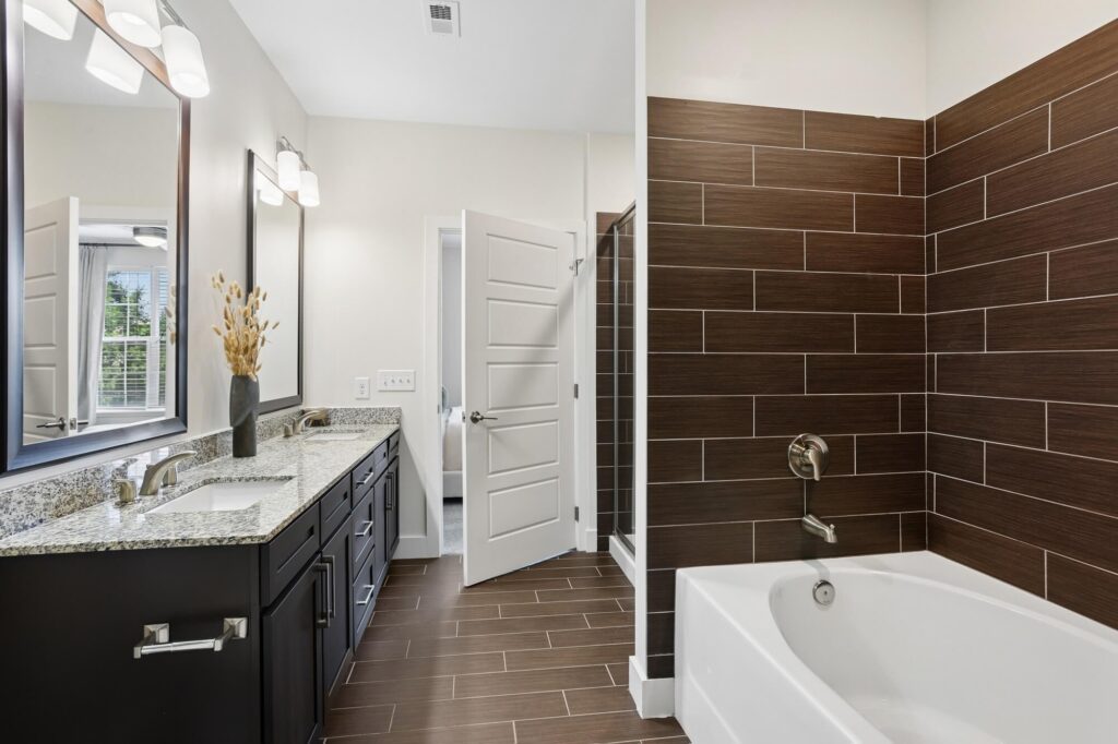Modern bathroom with a double-sink vanity, granite countertop, brown tile floor, white bathtub, and brown tiled shower. Theres a vase with dried plants on the counter and an open door in the background.