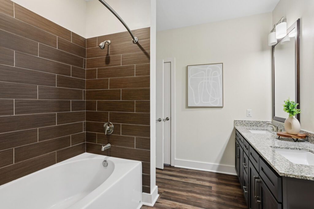 Modern bathroom with a bathtub and dark brown tile wall, double-sink vanity with granite countertop, large mirror, and decorative plant, featuring neutral-colored walls and wood flooring.