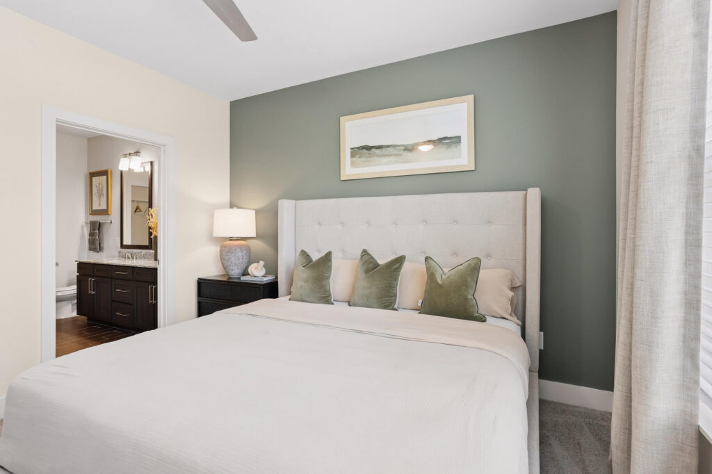 A neatly made bed with four green pillows sits against a green accent wall. A landscape painting hangs above the bed. A nightstand with a lamp is beside the bed. An open doorway leads to a bathroom.