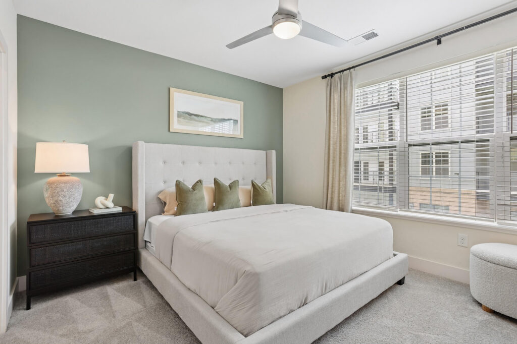 Modern bedroom with a large upholstered bed, four green pillows, a black nightstand with a lamp, a ceiling fan, green accent wall, and a window with blinds and beige curtains letting in natural light.