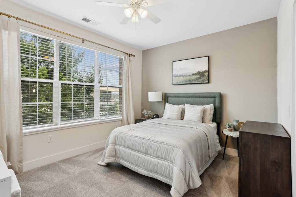 A bright bedroom with a large window, light beige walls, a green upholstered bed with white bedding, a nightstand with a lamp, a dresser, and a landscape painting above the bed.