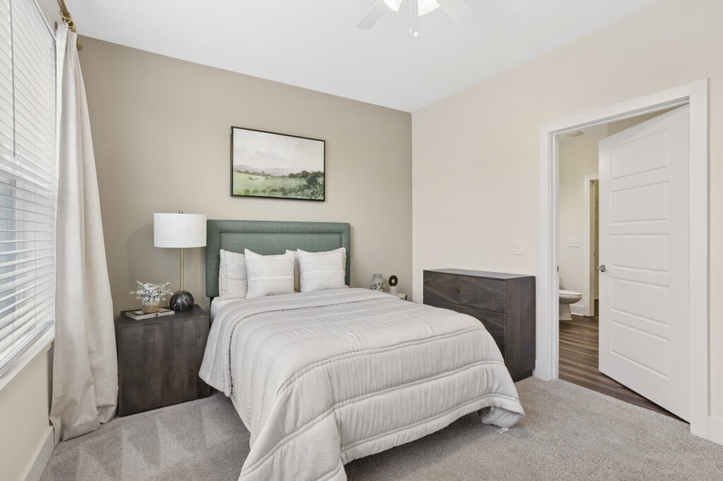 A tidy bedroom with a green upholstered bed, white bedding, two dark nightstands, a lamp, wall art above the bed, beige walls, carpeted floor, and an open door leading to a bathroom.