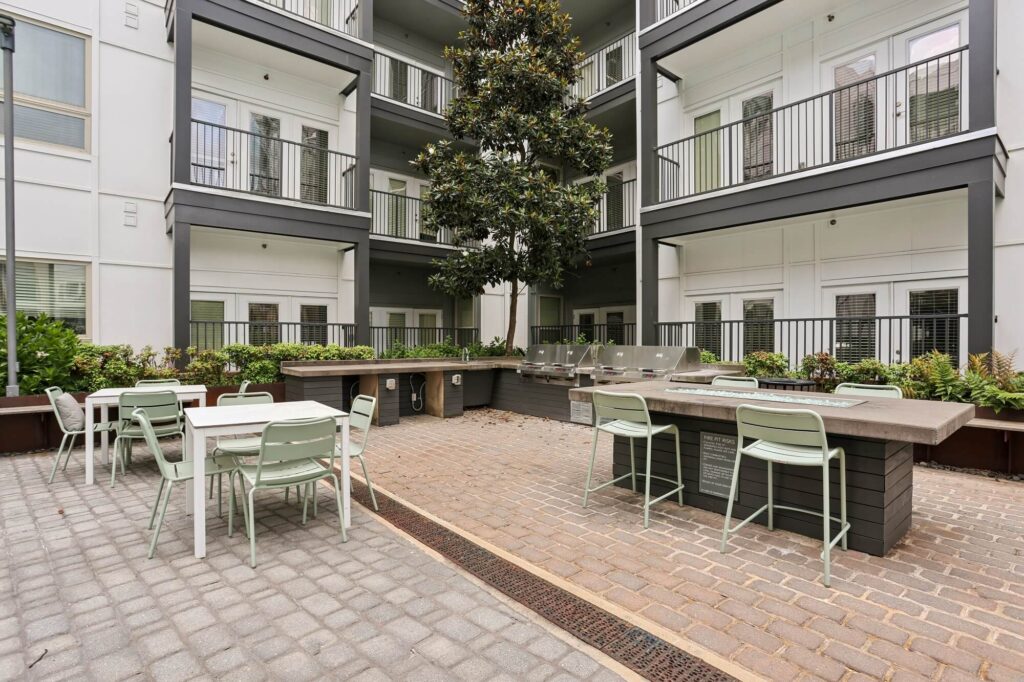 Modern apartment courtyard with outdoor seating, tables, and built-in grills. Surrounded by balconies and greenery, the space is designed for socializing and relaxation.