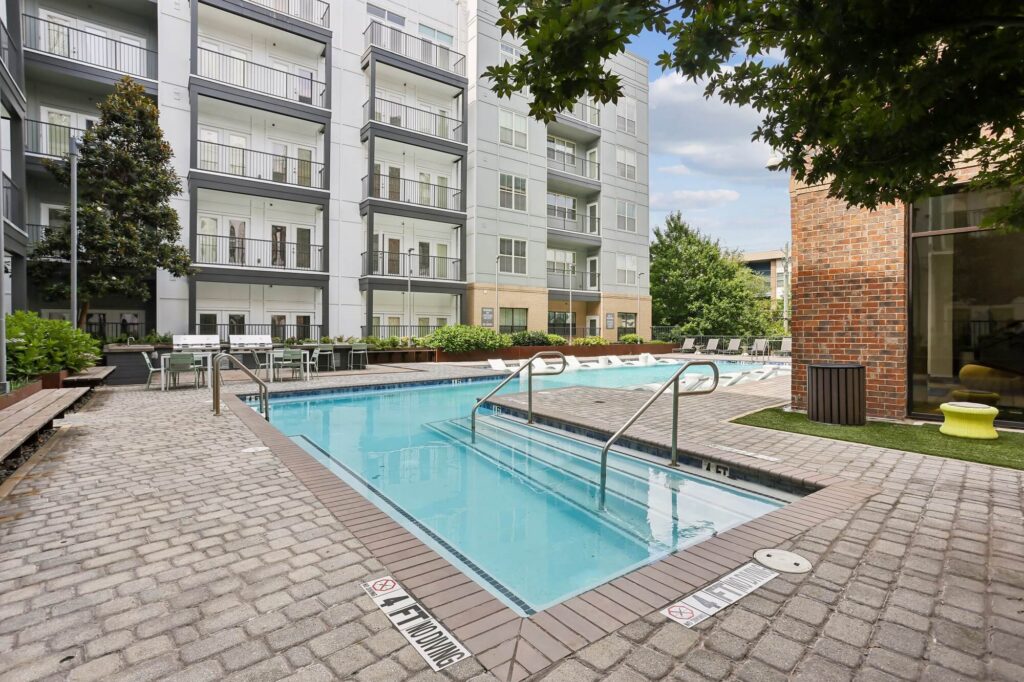 Outdoor pool area at an apartment complex with lounge chairs, potted plants, and a brick walkway. Multi-story buildings with balconies surround the pool. Trees and greenery add a relaxing atmosphere.