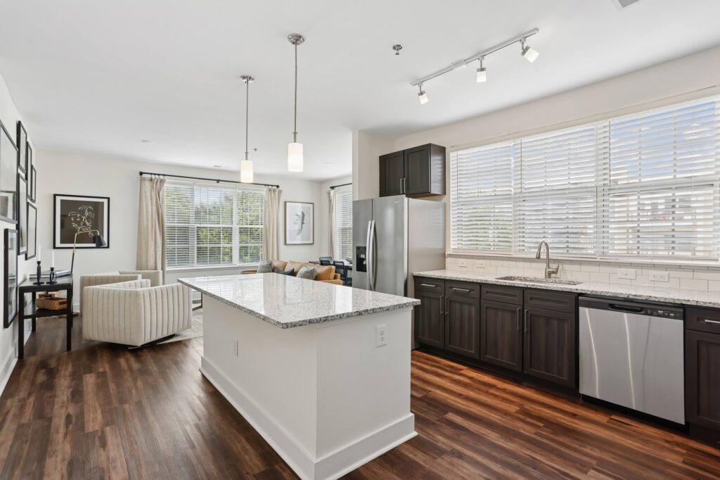 Modern kitchen with dark cabinets, stainless steel appliances, a large island with granite countertop, pendant lights, and wood flooring. Open layout leads to a bright living area with large windows and neutral decor.