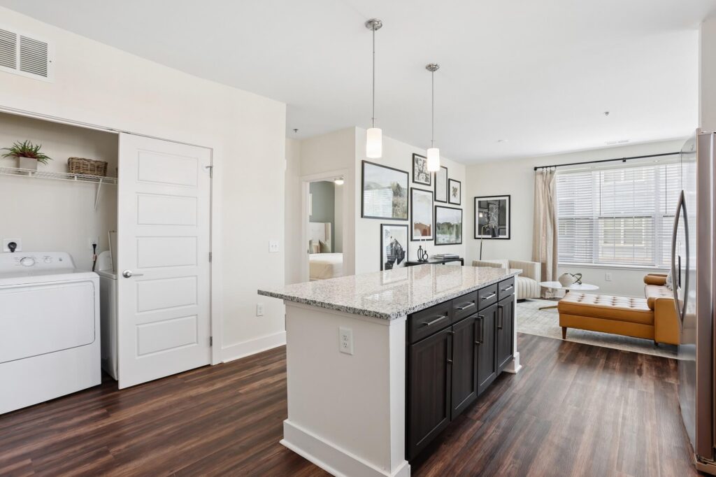 Modern apartment interior with an open kitchen, granite island, dark cabinets, pendant lights, laundry closet with washer and dryer, and a living room with a tan sofa and wall art by large windows.