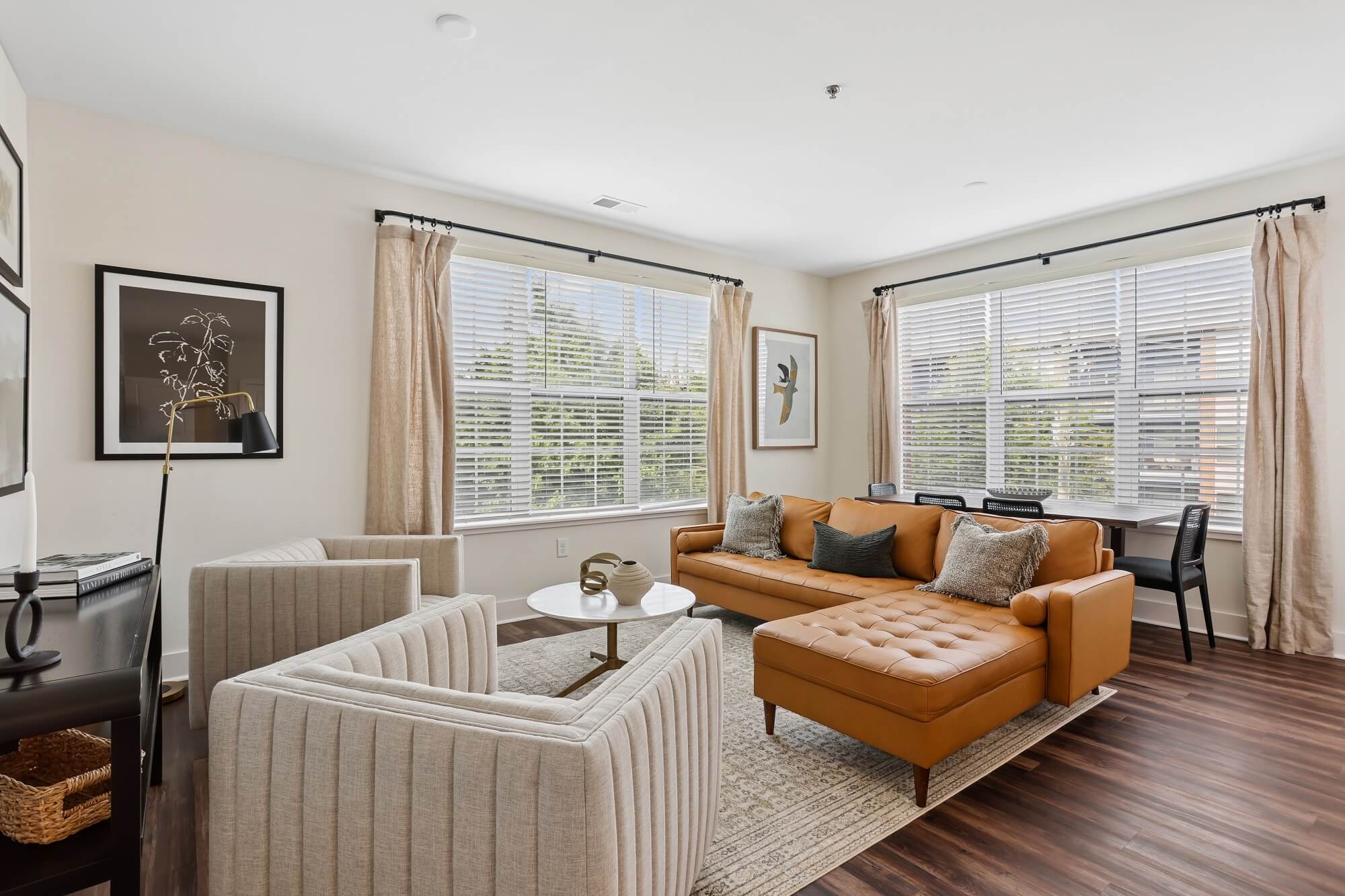 Modern living room with tan sofa, two beige armchairs, a round coffee table, desk with chair, large windows with beige curtains, and framed art on white walls. Dark wood floors and natural light fill the space.