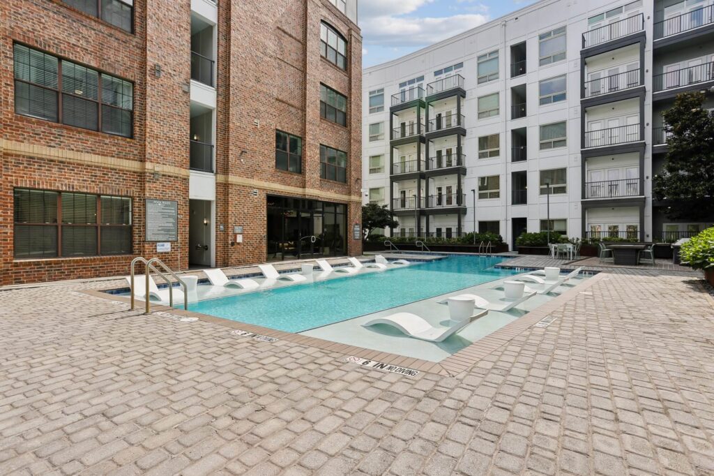 Modern apartment pool area with in-water lounge chairs, surrounded by brick and white apartment buildings, cobblestone deck, and greenery on a sunny day.