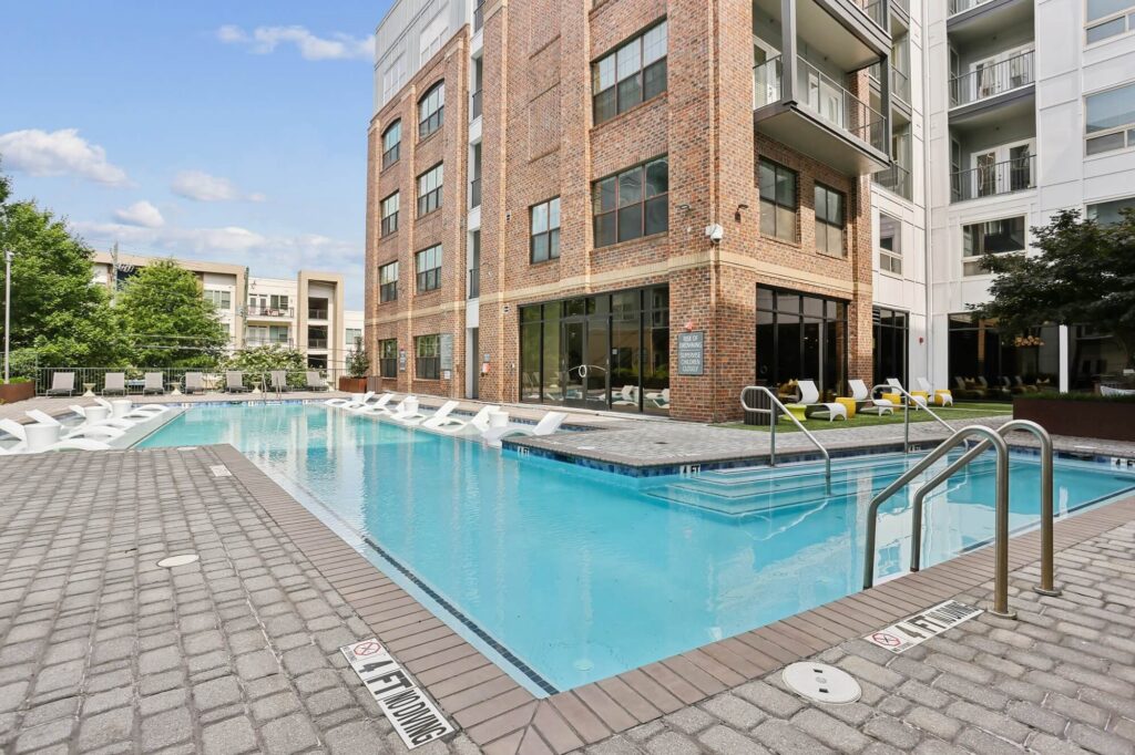 Rectangular outdoor pool beside a modern brick and glass apartment building, with lounge chairs partially in the water and more seating along the pool deck. Trees and other buildings are visible in the background.