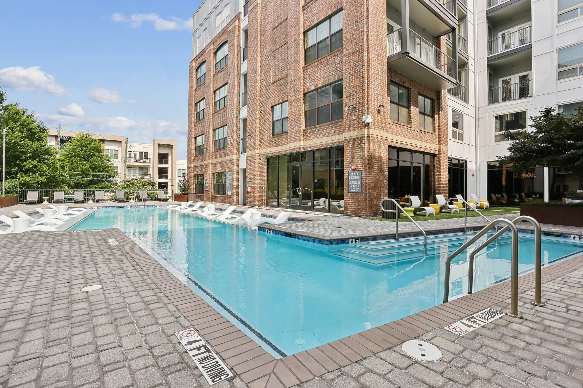 Rectangular outdoor pool beside a modern brick and glass apartment building, with lounge chairs partially in the water and more seating along the pool deck. Trees and other buildings are visible in the background.