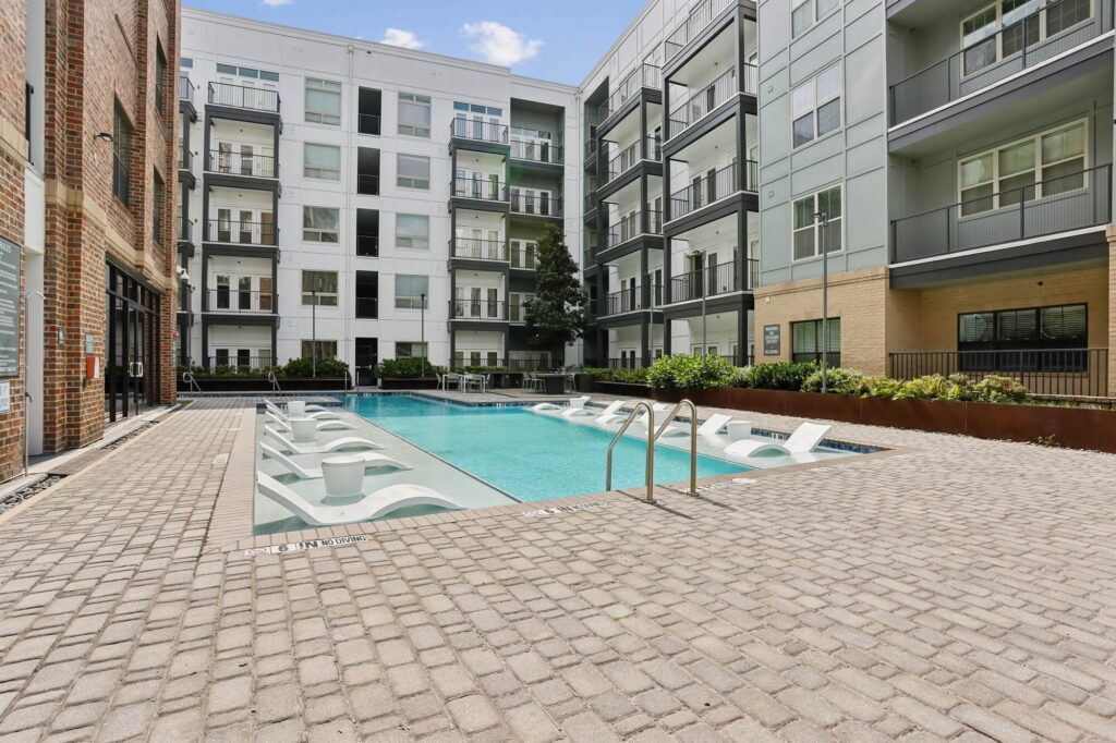 Rectangular outdoor pool with in-water lounge chairs, surrounded by a brick patio and modern multi-story apartment buildings, featuring balconies and greenery.