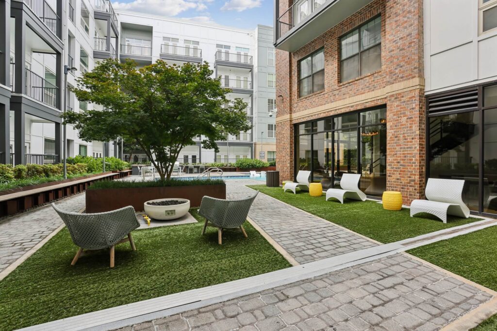 Modern apartment courtyard with outdoor seating, a tree in the center, artificial grass, brick walkways, and a pool in the background, surrounded by multi-story buildings and balconies.