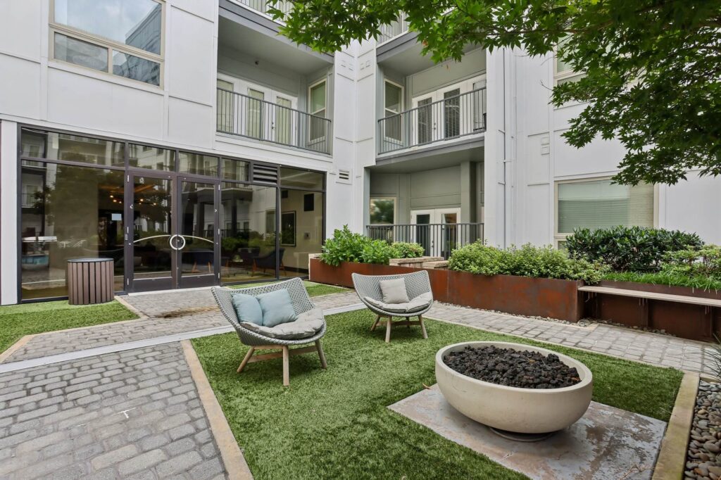 Modern apartment courtyard with two cushioned chairs, a round fire pit, artificial grass, pavers, large windows, and balconies. Green plants and a tree add natural elements to the quiet outdoor seating area.